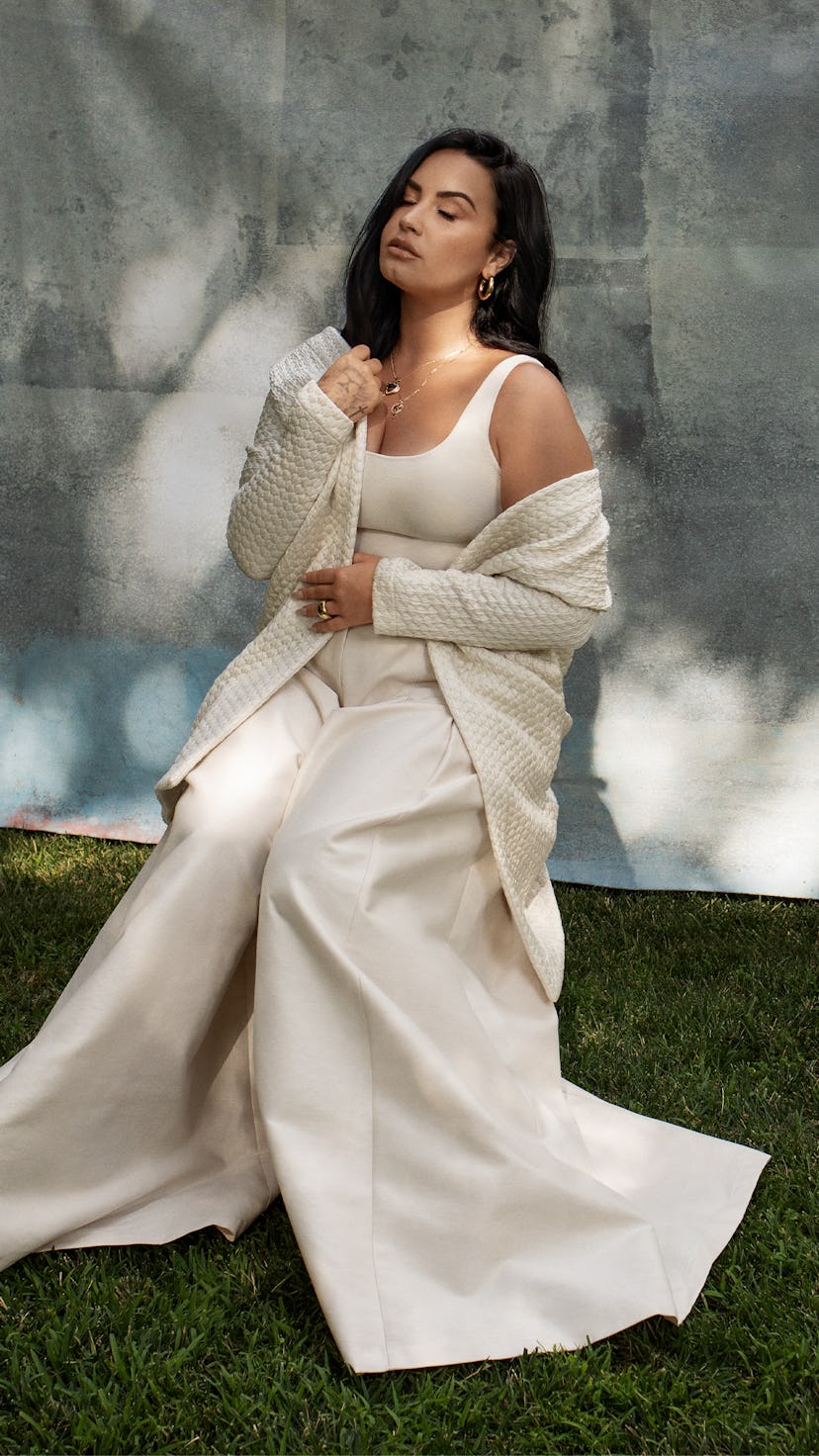 Demi sitting in baggy white pants and a white shirt with no sleeves on a chair in a garden