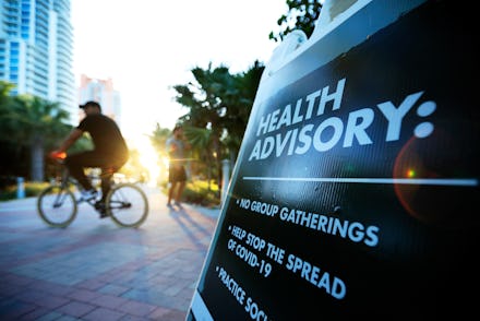 A sign on a street that says to stop group gatherings and Covid-19, with a bike driver in the blurre...