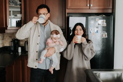 A mother and father sip coffee while holding a baby n their kitchen