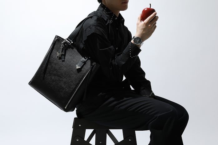 A man can be seen wearing a black shirt with black pants, holding a black bag and apple. His wrist p...