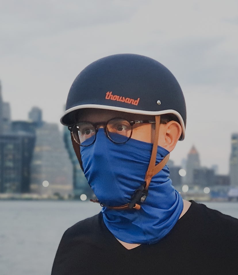 A man in a bicycle helmet with a buff over his mouth and nose standing in front of the Manhattan sky...