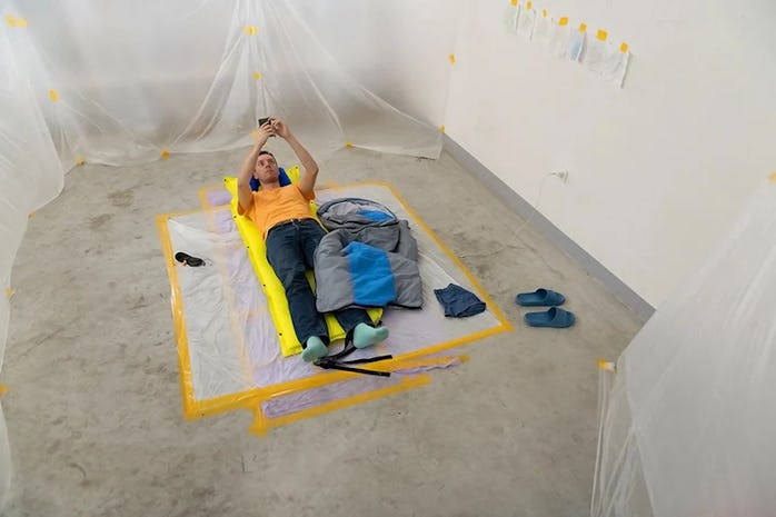 The artist Vincent Tanguy is shown in an orange shirt and jeans on the ground of a live cement house...