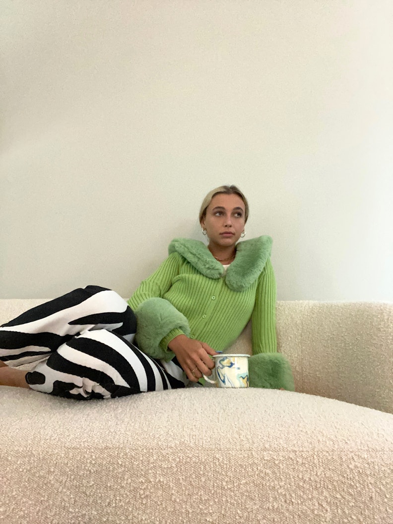 Internet personality Emma Chamberlain lying down on a bed wearing a green fur coat and black and whi...