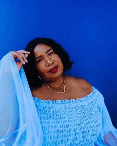 The influencer Kellie Brown posing in a light-blue tulle blouse and a thin necklace in front of a da...