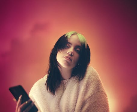 Billie Eilish looks at the camera, one hand holding a smartphone.