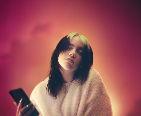 Billie Eilish looks at the camera, one hand holding a smartphone. 