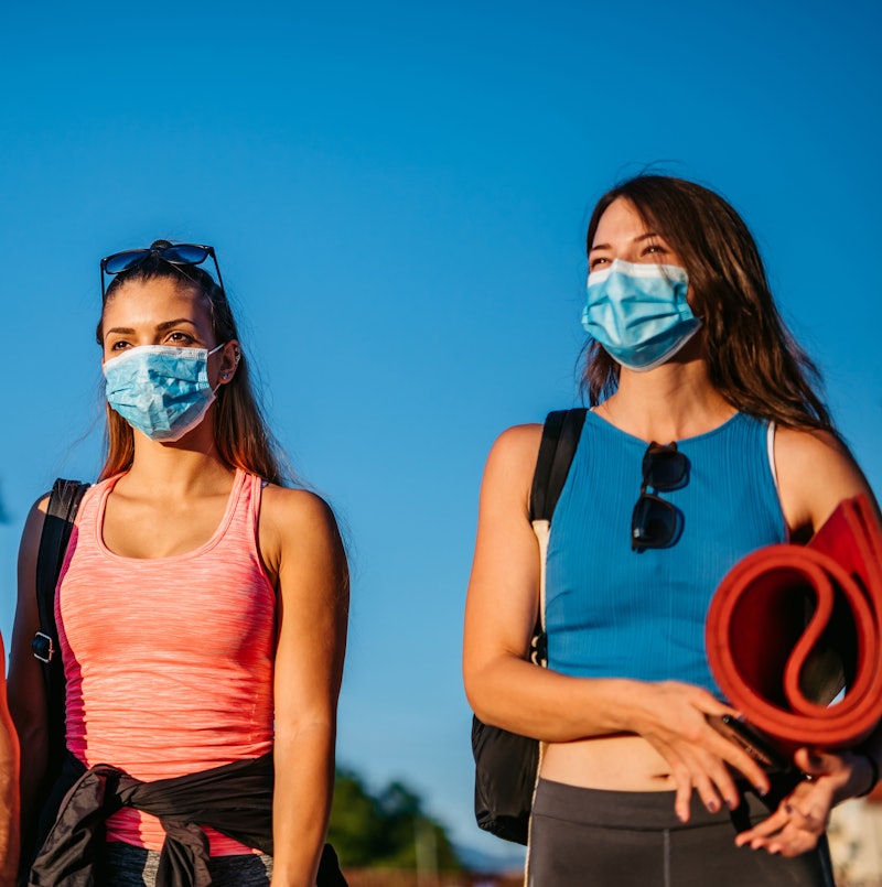 Two women wearing masks on their way to an outdoor yoga class. Are outdoor workout classes safe? we ...