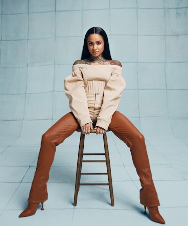 Kehlani sitting on a chair in white top and brown pants and boots by Burberry