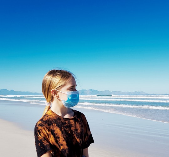 Young girl on beach wearing surgical mask, looking out at the ocean