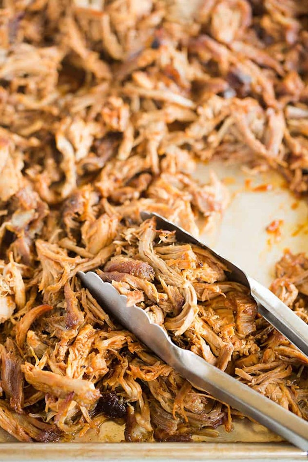 Closeup of tongs holding a portion of pulled pork, which was pulled from a larger sheet pan of pulle...