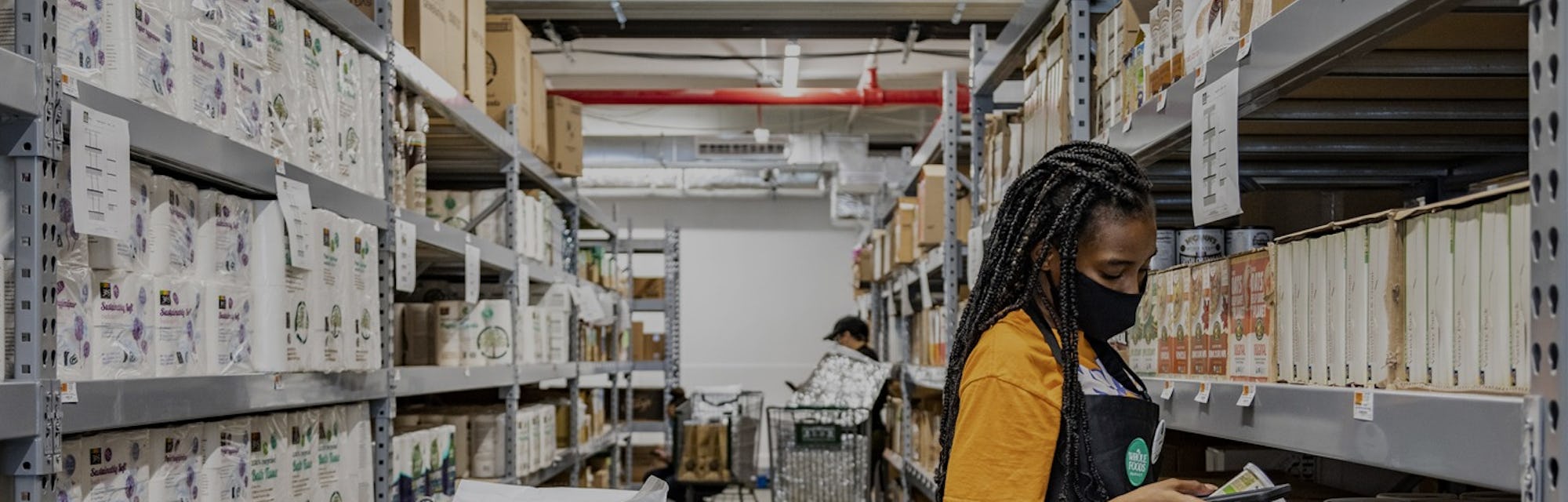 A young woman can be seen in a yellow shirt and black Whole Foods apron with a face mask on. She is ...