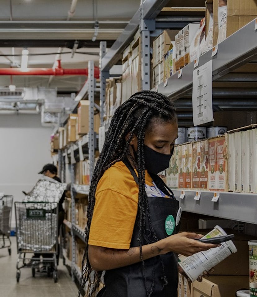 A young woman can be seen in a yellow shirt and black Whole Foods apron with a face mask on. She is ...