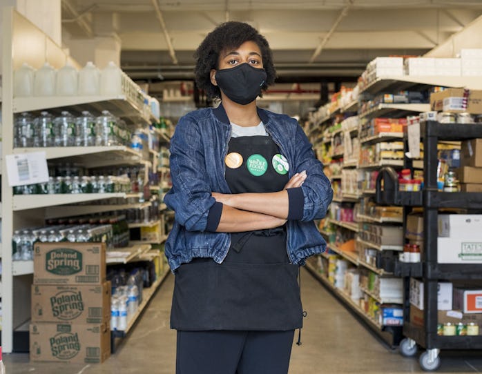 A young worker wearing a blue jacket and black Whole Foods apron is seen with her arms folded. Behin...