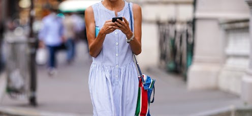 A woman walking down the street looking at her phone, wearing a light blue dress with her bag over h...