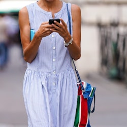 A woman walking down the street looking at her phone, wearing a light blue dress with her bag over h...