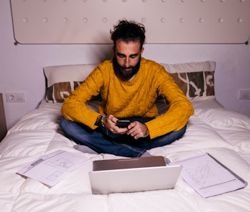 Young man working in bed at home with cell phone, laptop and documents.