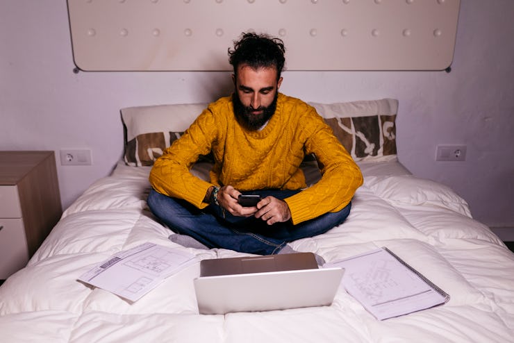 Young man working in bed at home with cell phone, laptop and documents.