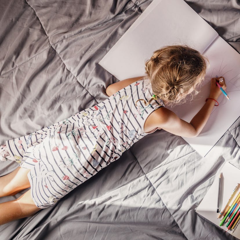 A girl lying on her stomach on crumpled bed sheets drawing in a large notebook