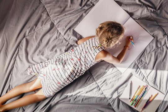 A girl lying on her stomach on crumpled bed sheets drawing in a large notebook