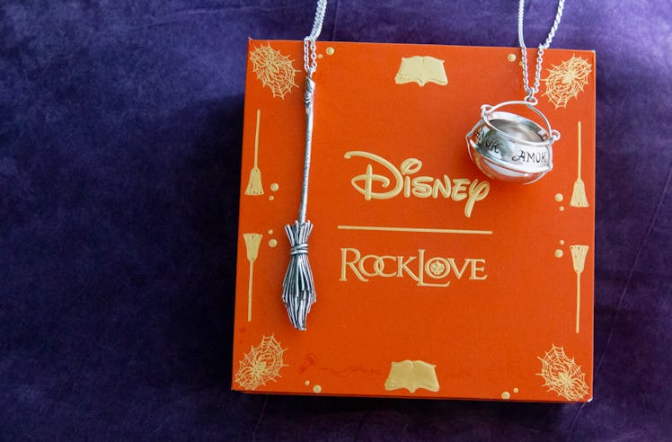 A broomstick and cauldron necklace from the Disney x RockLove 'Hocus Pocus' collection sits on an or...