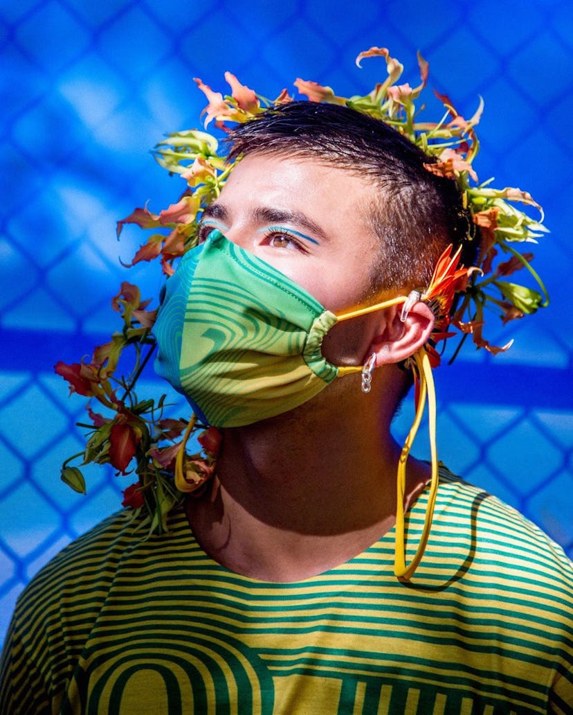 A model with flowers in his hair in front of a blue background with a fence, wearing a matching gree...