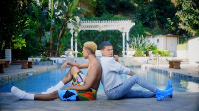 Rapper Kidd Kenn wearing a white shirt and jeans, sitting by the pool leaning on another person