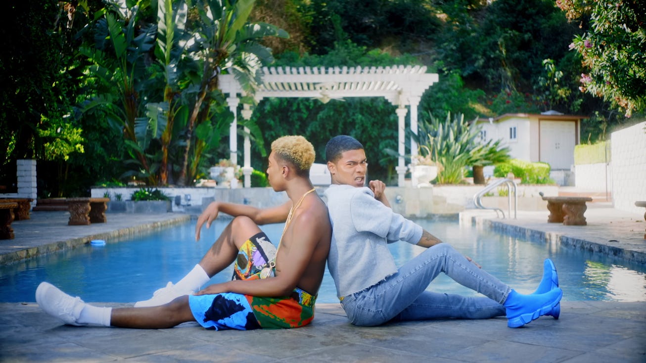 Rapper Kidd Kenn wearing a white shirt and jeans, sitting by the pool leaning on another person