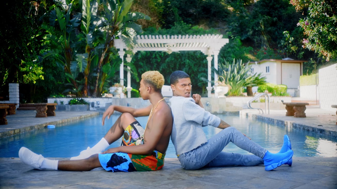 Rapper Kidd Kenn wearing a white shirt and jeans, sitting by the pool leaning on another person