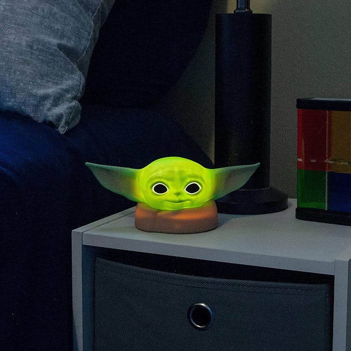 An image of a bed and nightstand with a soft looking Baby Yoda night light glowing atop the nightsta...