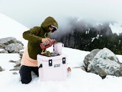 A woman pours a drink in a YETI tumbler on top of a YETI pink cooler. 