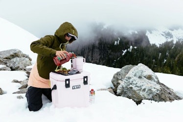 A woman pours a drink in a YETI tumbler on top of a YETI pink cooler. 