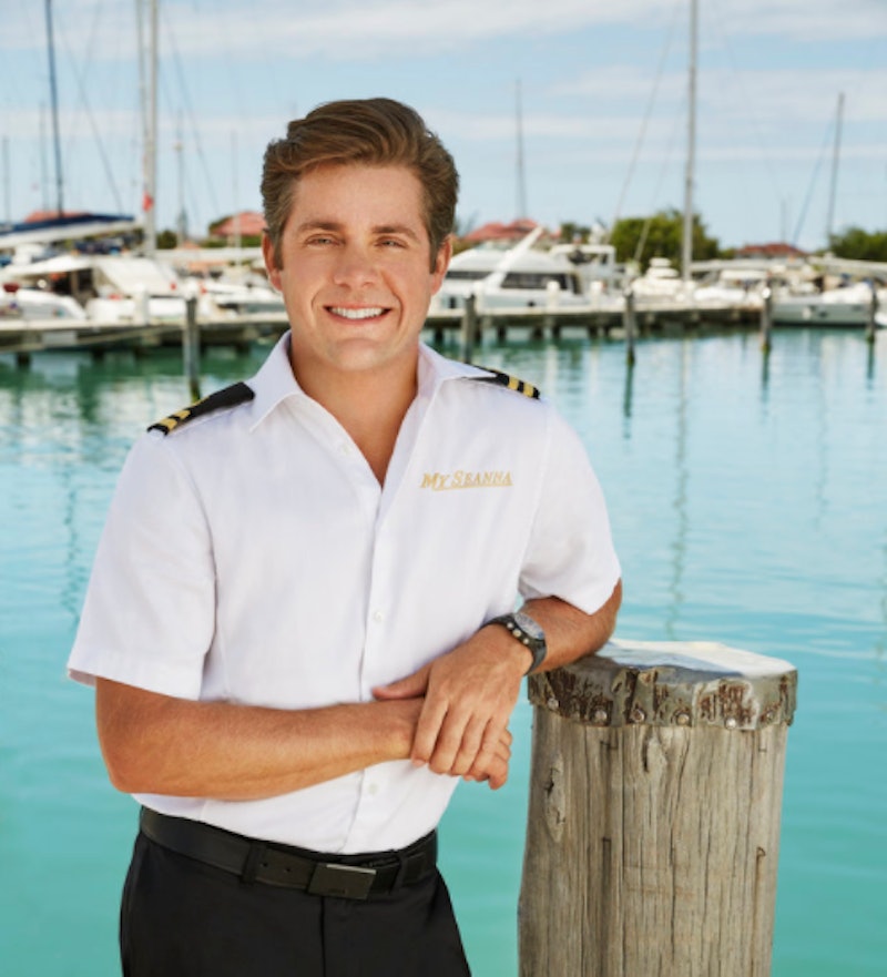 Eddie stands in his uniform in front of a marina with yachts moored on azure blue waters