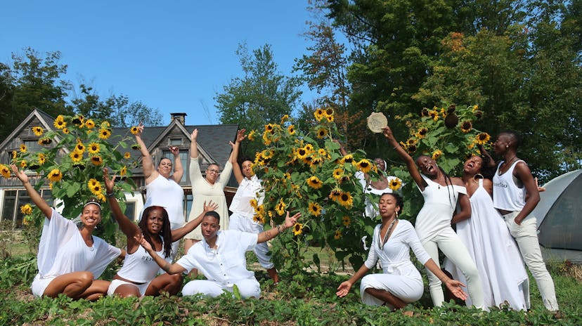 A group of women wearing all white, waving their hands up in the air, surrounded by sunflowers. 