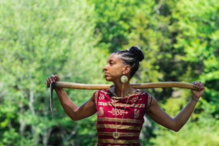 A woman with her hair braided, holding a spade on her shoulders, looking to her right.
