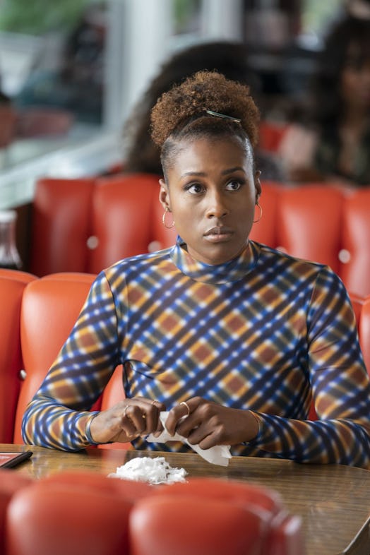 Issa Rae sitting in a red diner seat while wearing a blue-orange check turtleneck