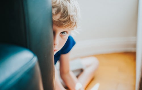 A little boy with curly blond hair peeks from behind a chair with an apprehensive expression.