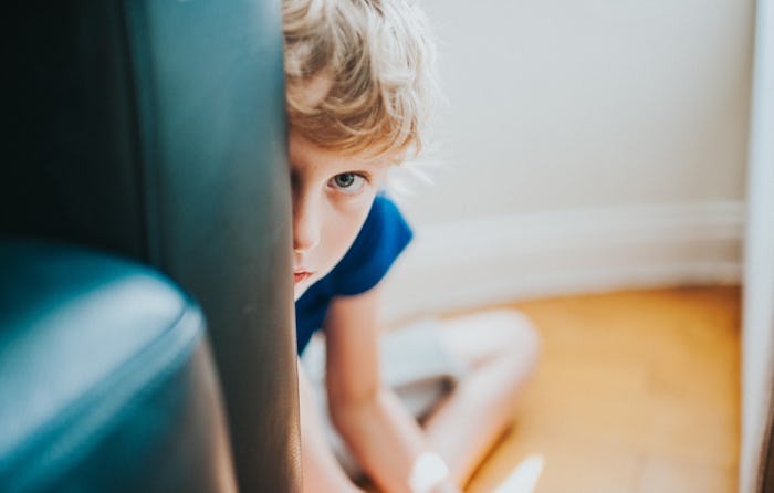A little boy with curly blond hair peeks from behind a chair with an apprehensive expression.