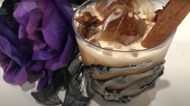 A halloween coffee drink sits on the table next to purple flowers in a skeleton hand cup.