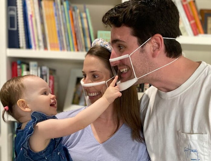 A mother and father are smiling brightly at their infant with transparent face masks on. The infant ...