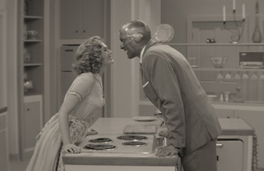 Elizabeth Olsen and Paul Bettany in 50s clothing leaning towards each other over a kitchen counter i...