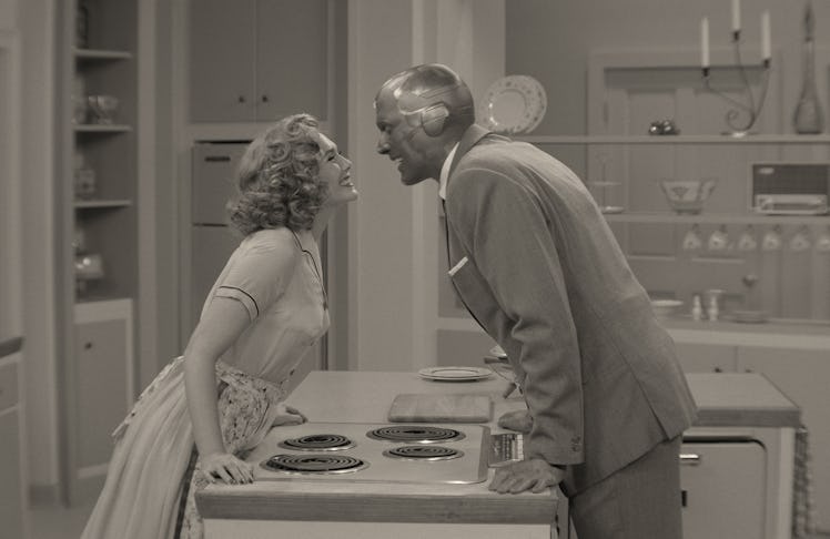 Elizabeth Olsen and Paul Bettany in 50s clothing leaning towards each other over a kitchen counter i...