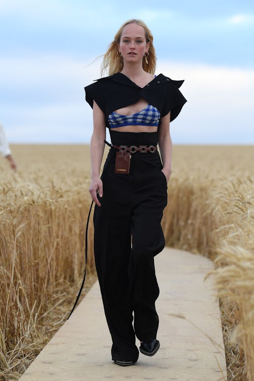 A model walking the runway in black pants and top over a blue gingham bralette and Jacquemus 'La Cei...