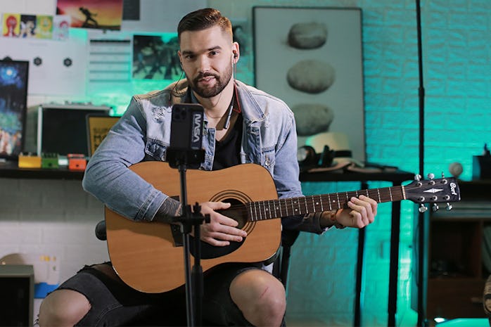 A musician holds a guitar while wearing Finalace headphones in front of a smartphone on a tripod