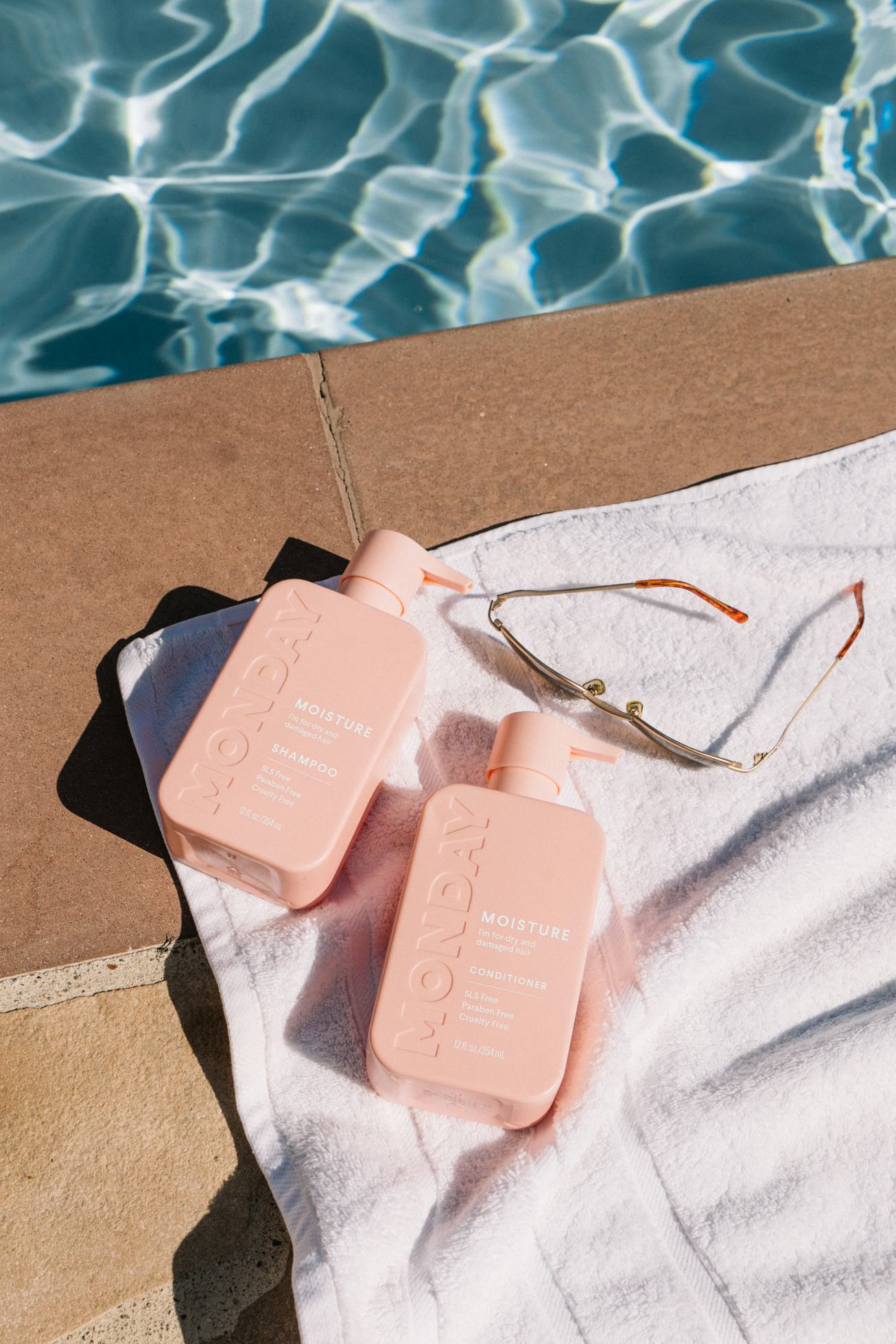 Two pink bottles of MONDAY hair care products on a towel by the pool.
