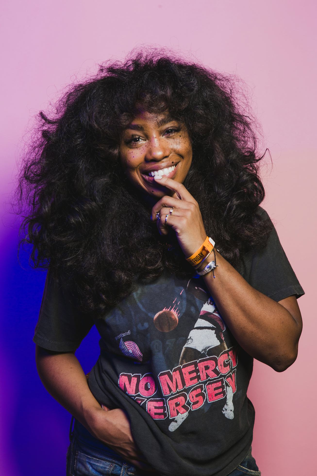 SZA in front of a pink wall with shoulder length curly hair and faux freckles.