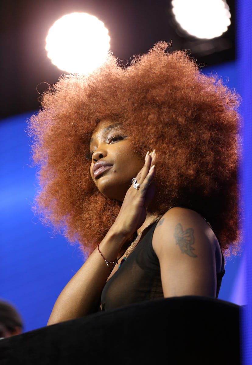 SZA in a black top wearing a large, round auburn afro wig.