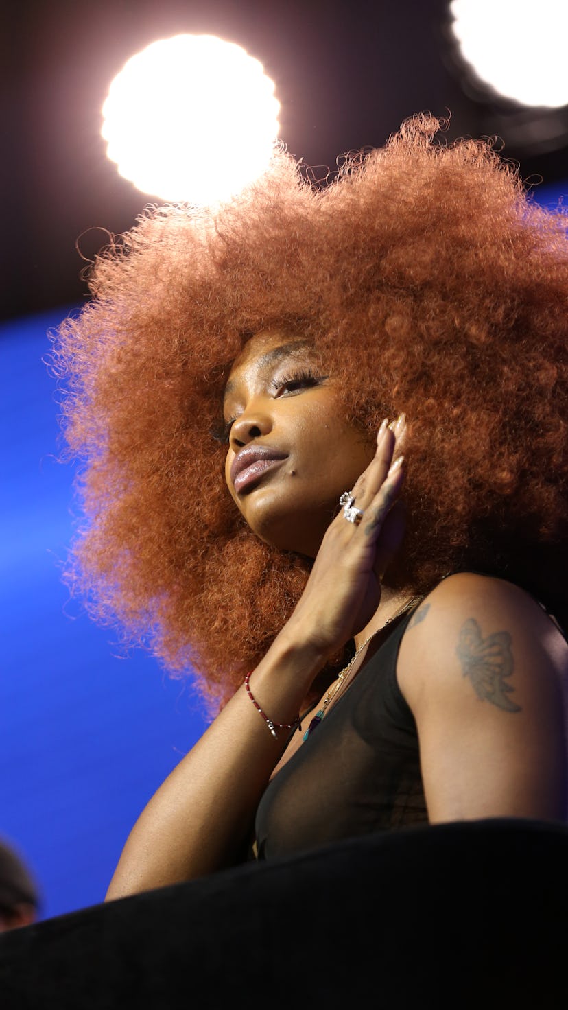 SZA in a black top wearing a large, round auburn afro wig.