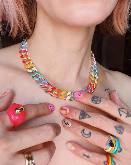 A close-up of a woman's neck with a rainbow chain necklace, and bold multi-colored rings on tattooed...