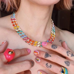 A close-up of a woman's neck with a rainbow chain necklace, and bold multi-colored rings on tattooed...