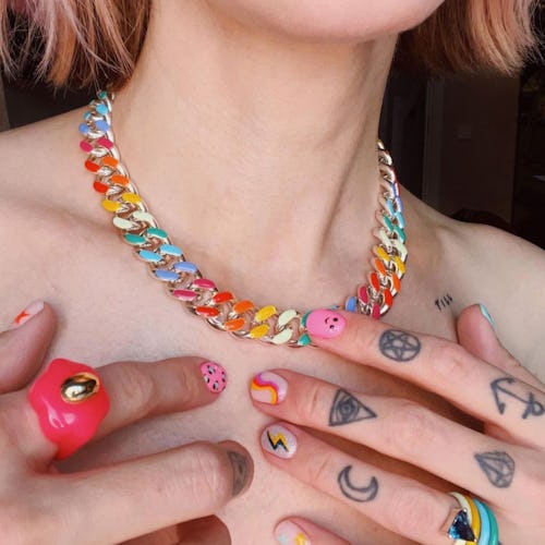 A close-up of a woman's neck with a rainbow chain necklace, and bold multi-colored rings on tattooed...